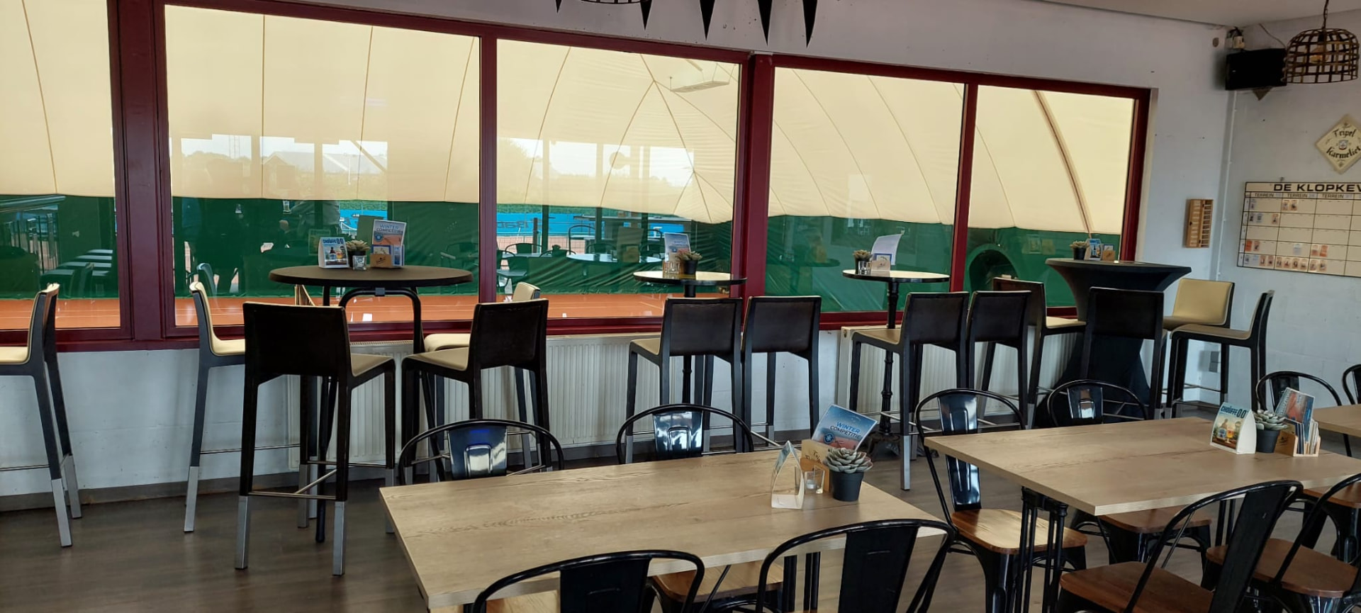 Bartafels en stoelen binnen in het café, langs de grote ramen met uitzicht op de tennisballon en de gravelvelden.