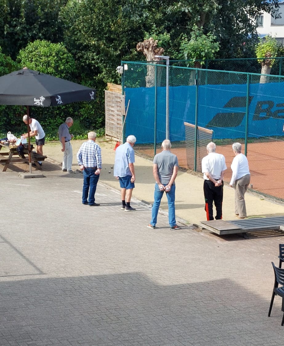 Een groep petanquers staat in de zon rond de petanquebaan naast het terras, met bomen en de tennisvelden erachter.