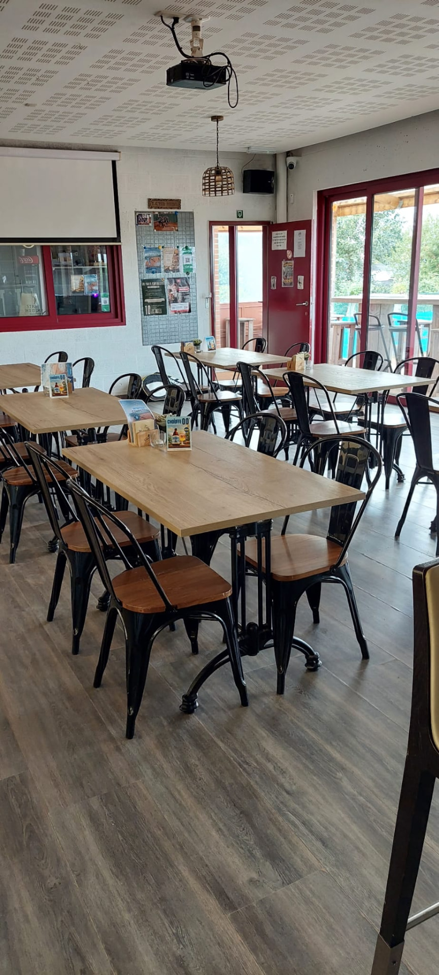 De zaal van het café met houten tafels, zwarte stoelen en een openstaande deur naar het terras.