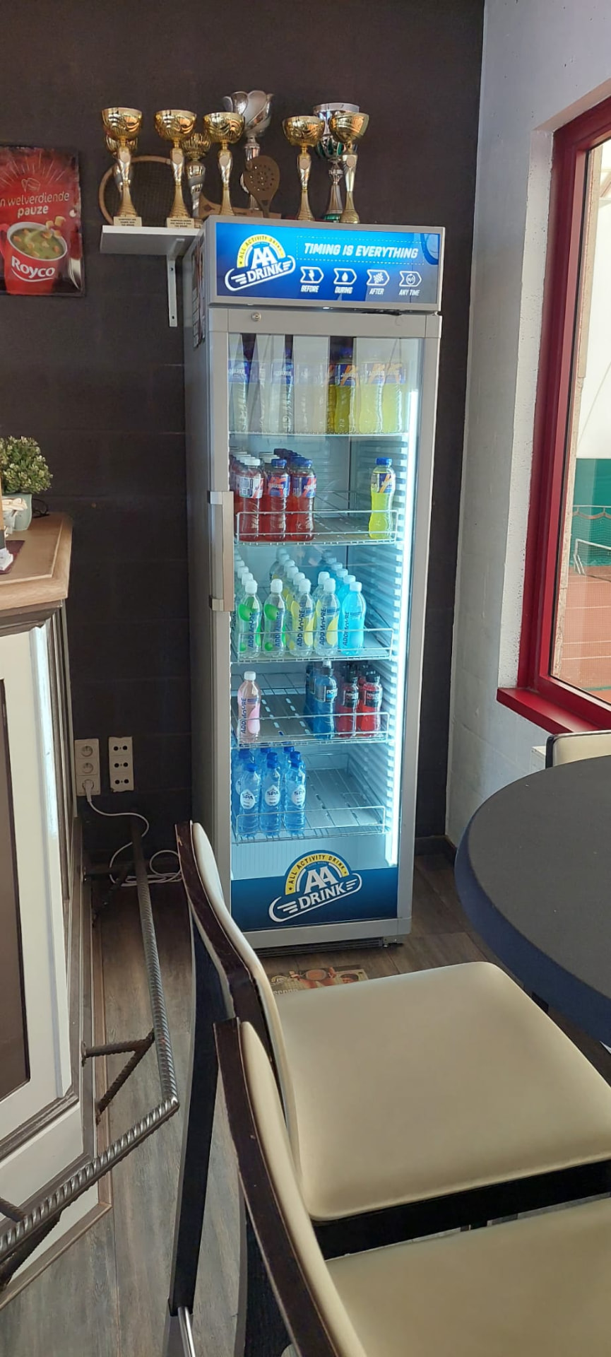 Een verlichte koelkast met flesjes sportdrank en water, met bekers en rackets op de kast erboven.